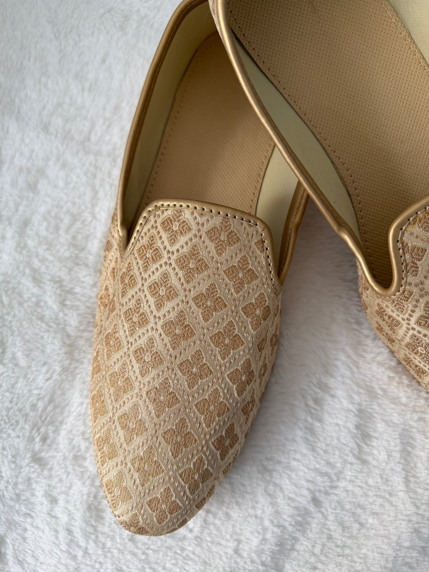Beige slip-on jutti with patterned toe cap on a white textured surface