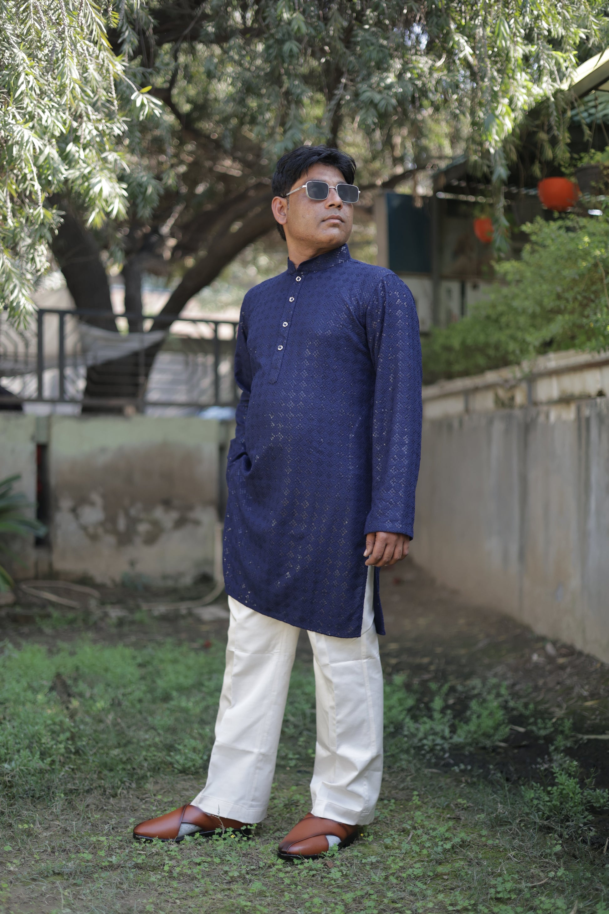 Person wearing a blue kurta and white pyjama standing outdoors with trees and a building in the background.