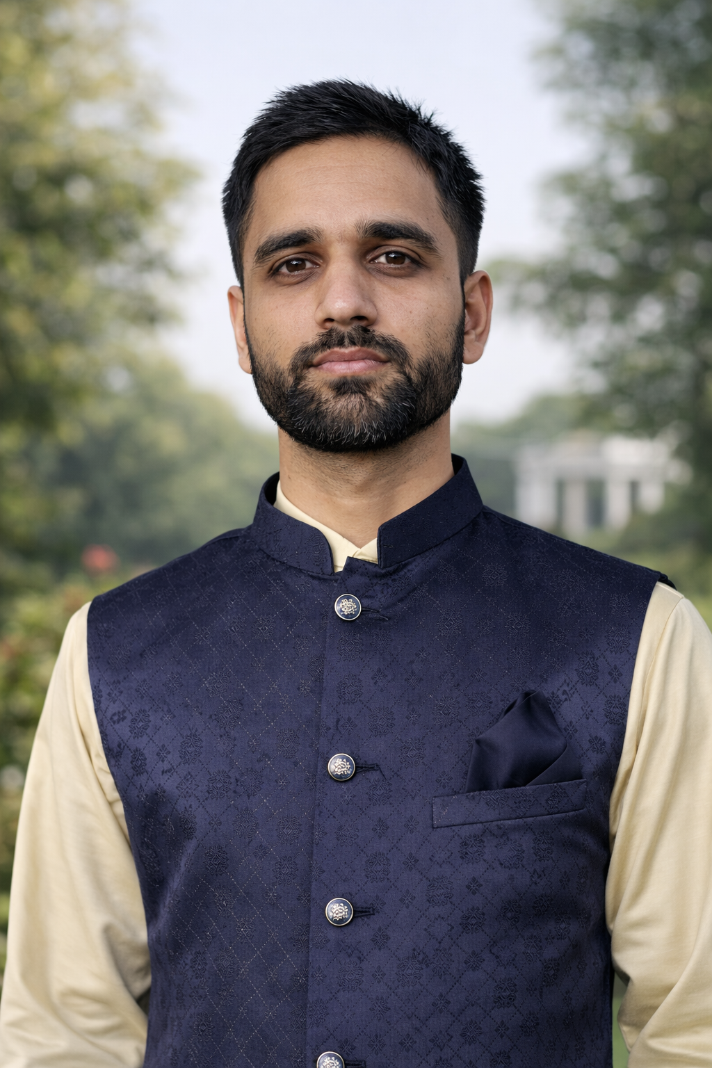 Man wearing a navy blue vest over a beige shirt with a blurred green background