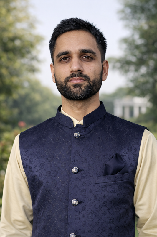 Man wearing a navy blue vest over a beige shirt with a blurred green background