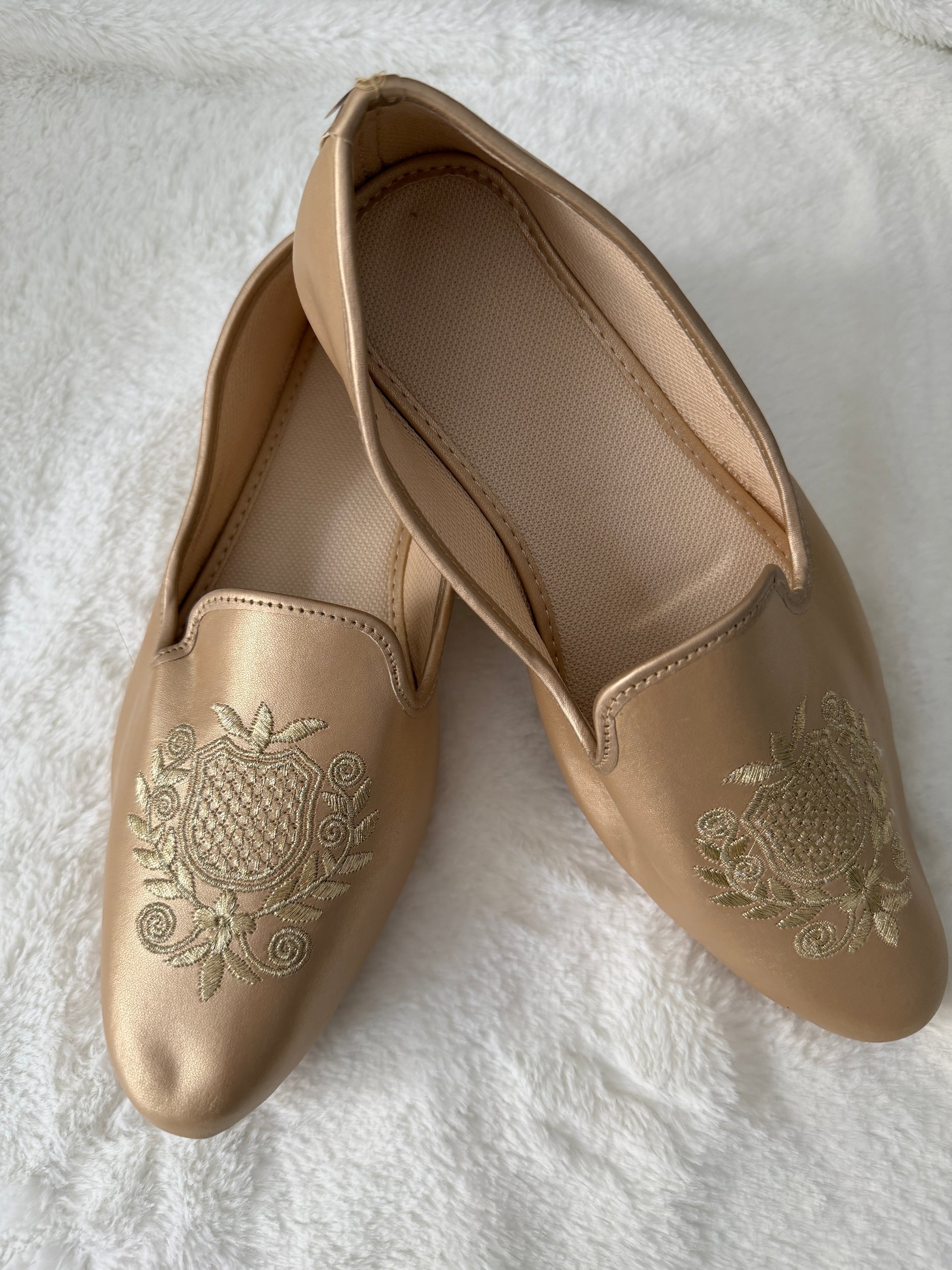 Pair of beige jutti with intricate gold embroidery on a white textured surface